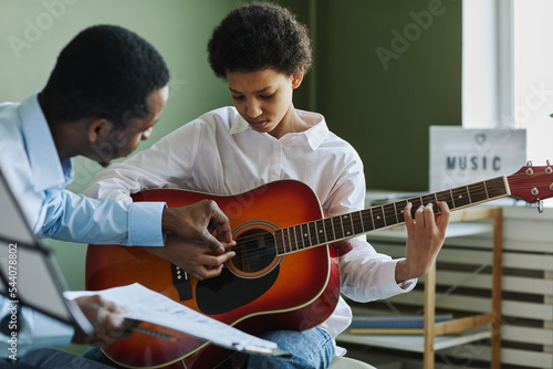 Fotografie Youthful diligent girl playing acoustic guitar while sitting in front of African