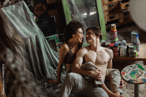 happy shirtless artist and african american woman in top sitting on floor near paints in art studio.