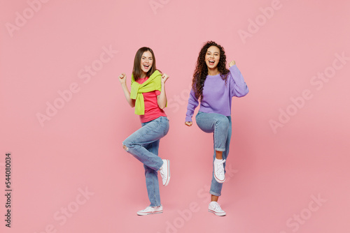 Full body young two friends overjoyed happy fun cool cheerful smiling women 20s wear green purple shirts together do winner gesture raise up leg isolated on pastel plain light pink color background.