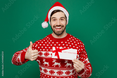 Merry young man wear red warm knitted sweater Santa hat posing hold gift certificate coupon voucher card for store show thumb up isolated on plain dark green background New Year 2023 holiday concept.