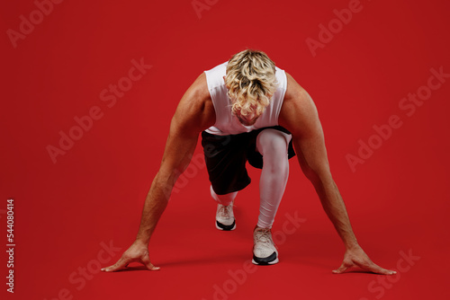 Full body young strong sporty toned sportsman man wear white clothes spend time in home gym stand at low start going to run marathon isolated on plain red background. Workout sport fit body concept.