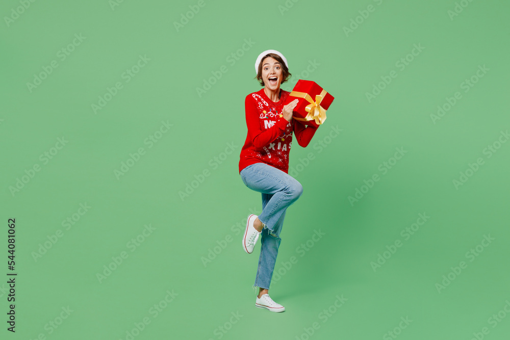 Full body merry young woman wear knitted xmas sweater Santa hat posing hold red present box with gift ribbon bow isolated on plain pastel light green background. Happy New Year 2023 holiday concept.
