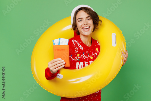Traveler woman in xmas sweater Santa hat hold swim ring passport ticket isolated on plain green background Tourist travel abroad in free time rest getaway Air flight trip Happy New Year 2023 concept. #544081020