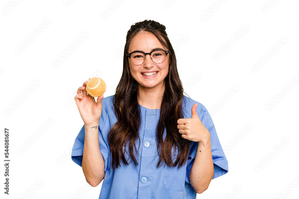 Young caucasian physiotherapist woman holding a tennis ball isolated isolated on green chroma background smiling and raising thumb up Young caucasian physiotherapist woman holding a tennis ball isolated isolated on green chroma background smiling and raising thumb up