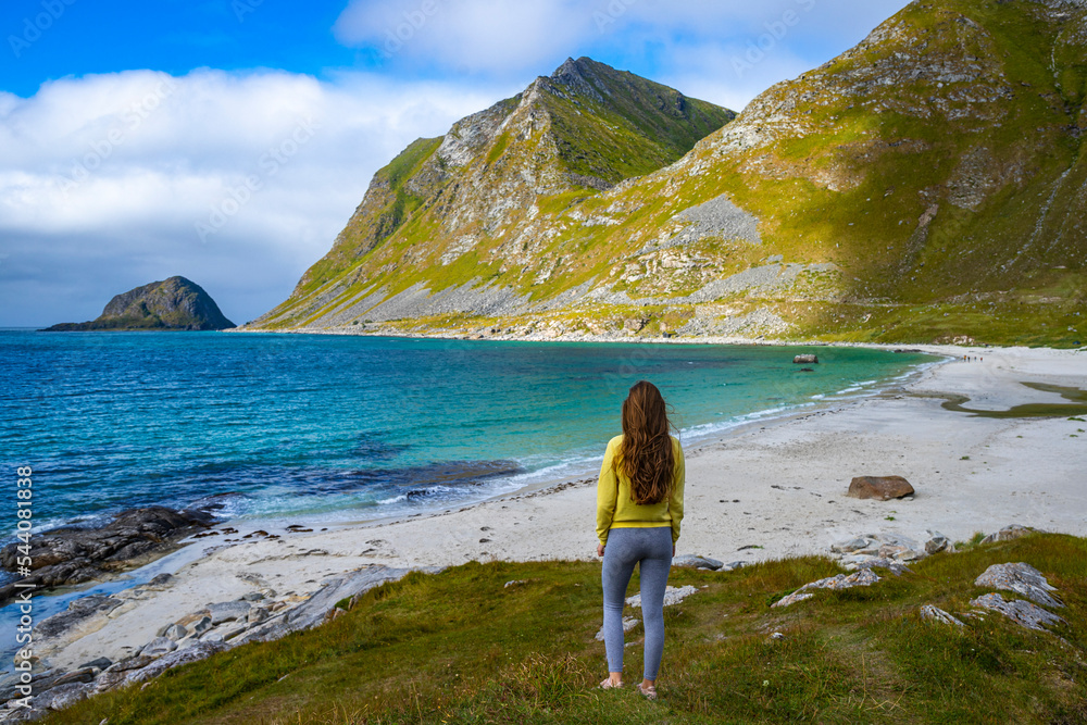 beautiful long-haired girl stands on a hill admiring the panorama of the famous haukland beach on lofoten islands in norway; relaxing on a paradise beach in norway