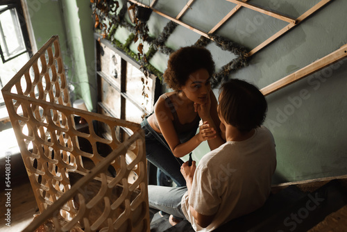 high angle view of young artist sitting on stair in workshop and touching face of sexy african american woman.