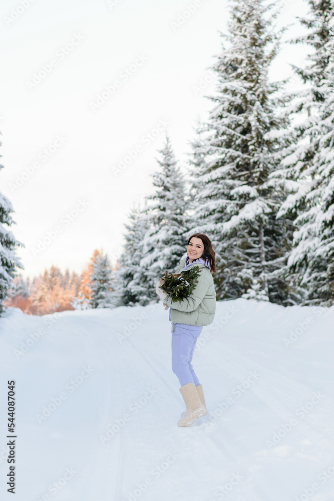 Happy young dark-haired woman in a down jacket and a lilac suit walking in the winter forest with fir branches in her hands.Winter outdoor activities,New Year,Christmas and eco-friendly concept.
