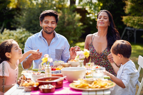 Religious Family Saying Prayers Or Grace Before Eating Outdoor Meal In Garden