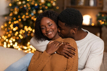 Smiling young black husband hug and kiss wife, enjoy holiday together in living room interior with Christmas tree
