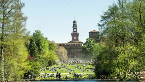 Palazzo del Arte and tourists on lawn in historical center of Milan on windy sunny day. Parch Sempion with lush greenery and river hyper lapse