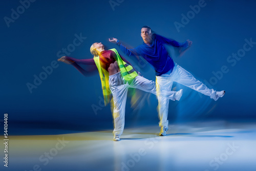 Portrait of young man and woman dancing isolated over dark blue background with mixed lights. Synchronic photo