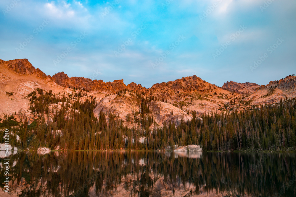 Alpine Lake, located in Idaho’s Sawtooth Wilderness, seen on a summer morning at dawn, with orange sunlight illuminating Packrat and Monte Verita Peaks. The scene is reflected in the tranquil waters.
