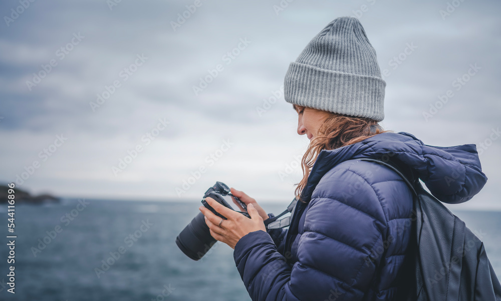 A young cheerful woman in a scarf jacket and a hat, a traveler professional photographer with a camera in her hands on the shore of the winter sea