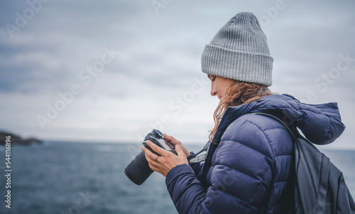 Obraz na płótnie A young cheerful woman in a scarf jacket and a hat, a traveler professional photographer with a camera in her hands on the shore of the winter sea w salonie