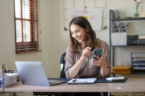 Smiling and charming Asian woman sit at her desk with a smartphone playing social media applications and transacting at her desk.