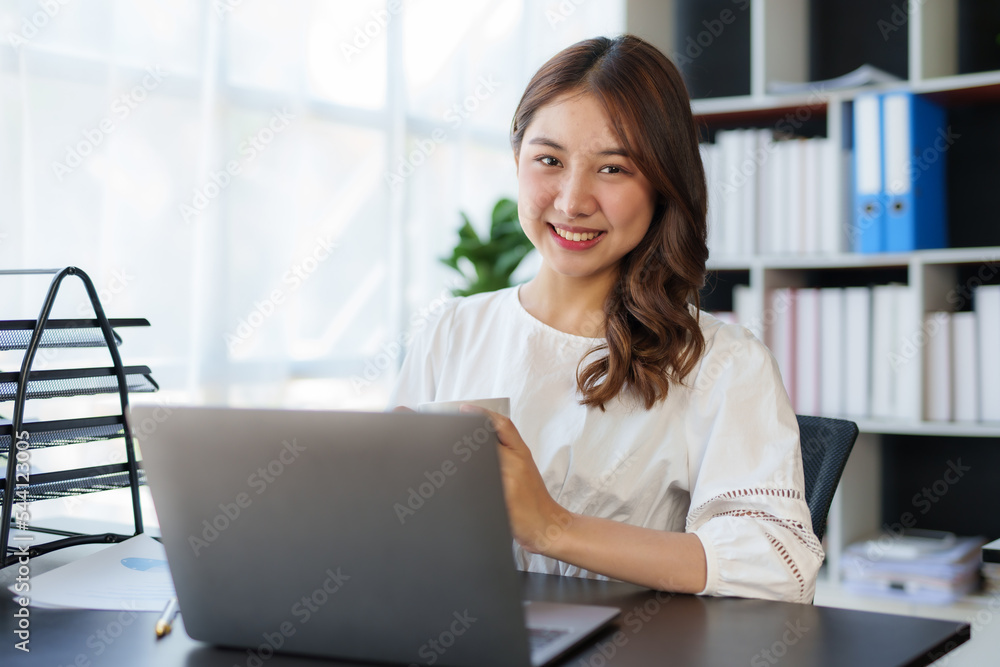 Beautiful Asian businesswoman sitting on her laptop happily working on her laptop and drinking coffee with a bright smile in the office and looking at the camera. Beautiful Asian businesswoman sitting on her laptop happily working on her laptop and drinking coffee with a bright smile in the office and looking at the camera.