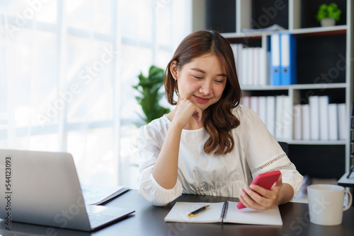Photos Smiling and charming Asian woman sit at her desk with a smartphone playing social media applications and transacting at her desk