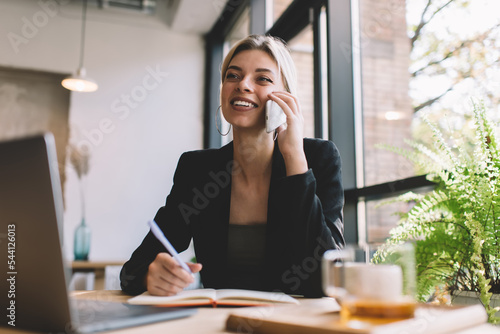 Cheerful female entrepreneur with laptop and textbook using cellphone device for calling and talking about business ideas, happy Caucasian woman discussing mobility communication while phoning