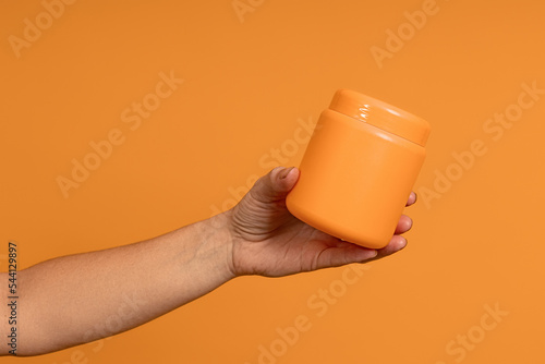 a female hand holds out an opaque plastic yellow jar on a yellow background
