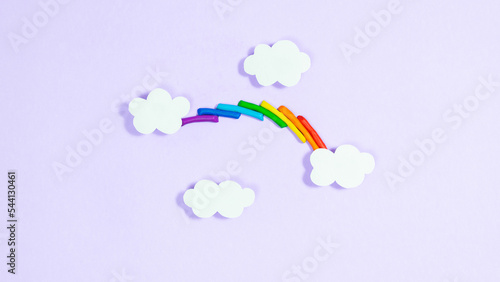 Colorful plasticine rainbow between white paper clouds. Minimalism, copy space. Template for design and text. Light purple background.