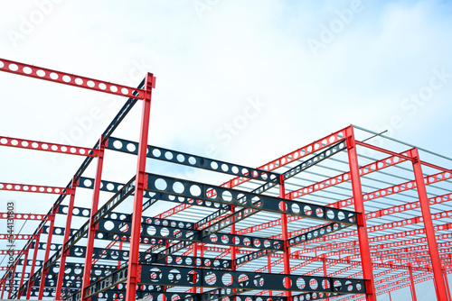 Red and black castellated beam metal structure of large industrial building in construction site against white cloudy sky background