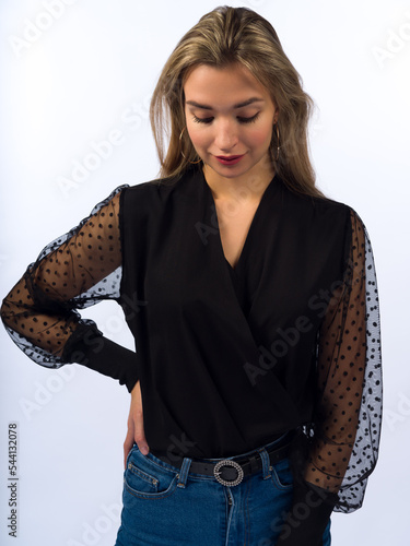 A beautiful, young, slender girl with long blonde hair on a white background in blue jeans and a stylish black blouse with transparent sleeves. Close-up