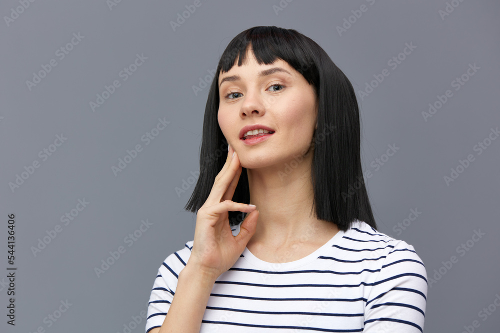 a sweet, attractive woman stands on a gray background in a white striped T-shirt and smiling pleasantly holds her fingers near her face in a relaxed gesture. Horizontal photo a sweet, attractive woman stands on a gray background in a white striped T-shirt and smiling pleasantly holds her fingers near her face in a relaxed gesture. Horizontal photo