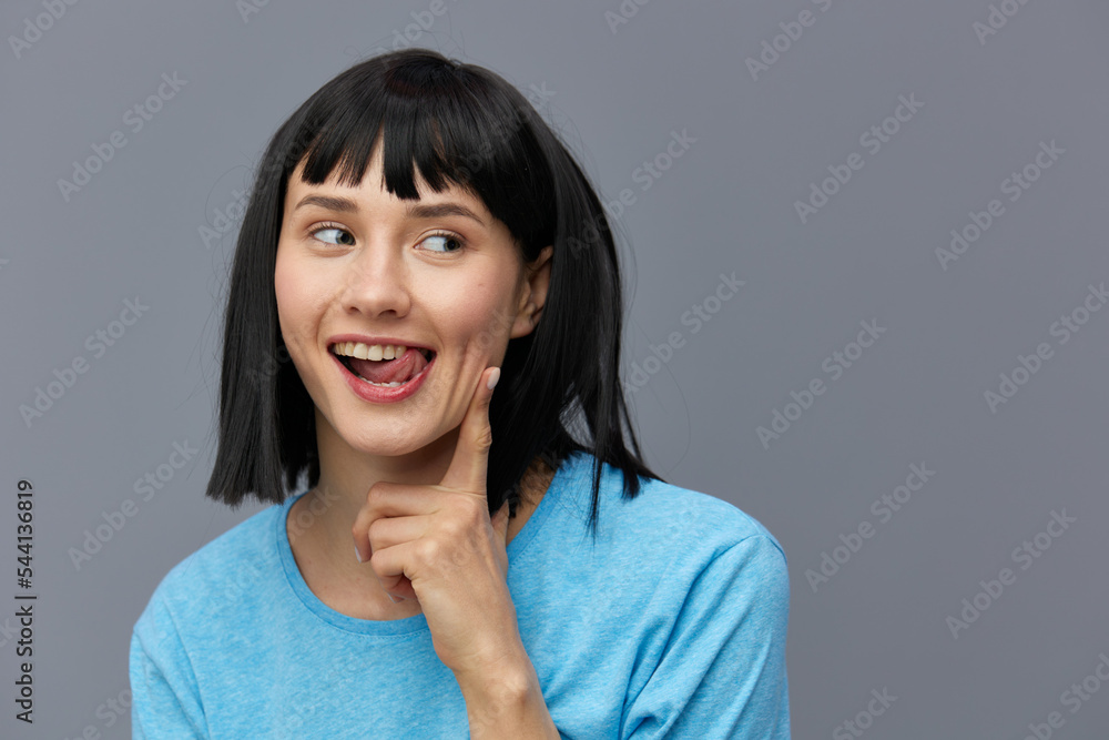 a beautiful, attractive, happy woman poses standing on a gray background in a blue T-shirt and happily looking at the camera with her index finger touching her face. horizontal studio photo a beautiful, attractive, happy woman poses standing on a gray background in a blue T-shirt and happily looking at the camera with her index finger touching her face. horizontal studio photo