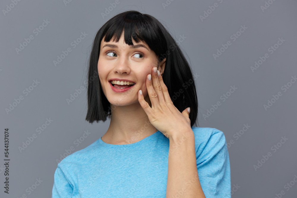 a beautiful, attractive, happy woman poses standing on a gray background in a blueT-shirt and happily looking at the camera with her index finger touching her face. horizontal studio photo a beautiful, attractive, happy woman poses standing on a gray background in a blueT-shirt and happily looking at the camera with her index finger touching her face. horizontal studio photo