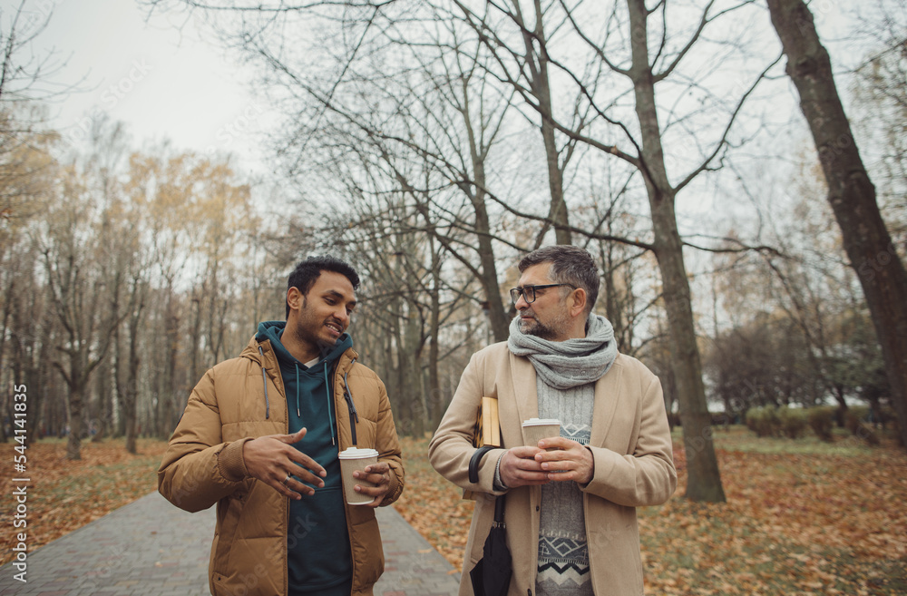 Friends, a senior and a young man walking and talking and drinking coffee together in the autumn park..