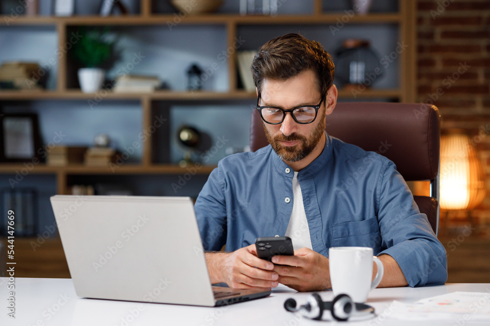 Obraz premium Handsome male businessman manager working behind a laptop in a stylish home office. Remote work at home. Portrait of a successful freelancer.