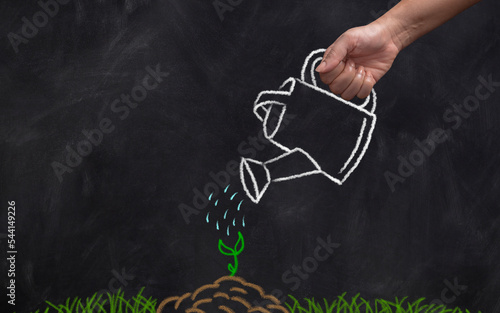 Close-up of hand on black board holding watering can drawn with chalk over green sprout.