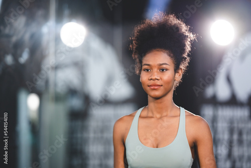 Portrait of beautiful and fit young African female with curly hair athlete training in gym clothes standing leaning towards machine for exercise while smiling and looking at camera