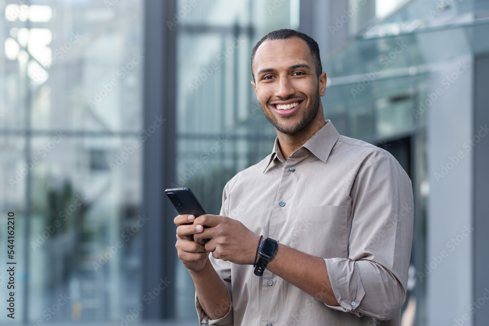 Fototapeta premium Portrait of African American businessman, man holding phone smiling and looking at camera, office worker in shirt outside modern office building on lunch break.