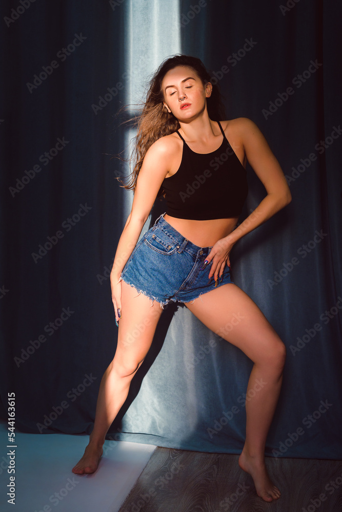 Full length portrait of an attractive brunette woman with long curly hair in the studio at sunset light. Model in black tank top and denim shorts barefoot indoors. beautiful light and shadow Full length portrait of an attractive brunette woman with long curly hair in the studio at sunset light. Model in black tank top and denim shorts barefoot indoors. beautiful light and shadow