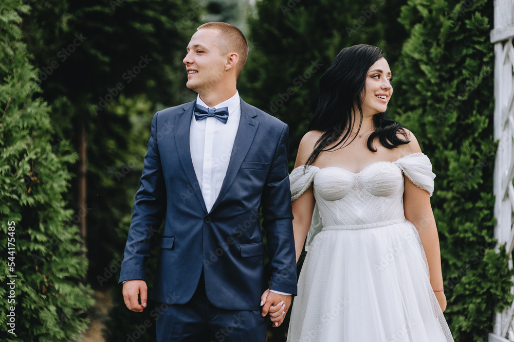 A fashionable groom in a blue suit and a beautiful, smiling, curly brunette bride in a white dress are standing near a wooden arch, holding hands, in a park in nature. wedding photography.