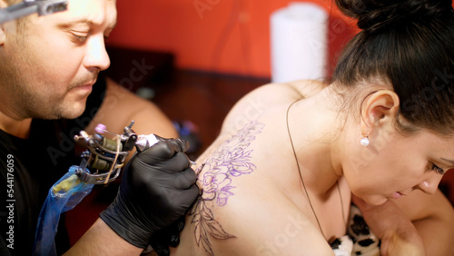 in a tattoo salon , a specialist is doing a tattoo on woman's back, a floral ornament. a man works in special gloves, on special equipment. drawing is done with black paint. High quality photo photo