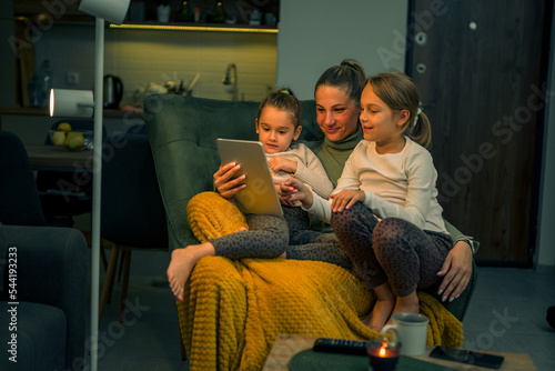 Mother and daughters are comfortable on an armchair looking at videos on a tablet, they are happy