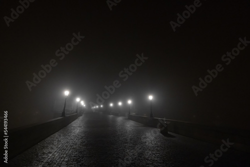 Charles or Karlov bridge in Prague in early morning hours with a lot of fog and haze, with visible lights and no people. Spooky scary bridge in prague.