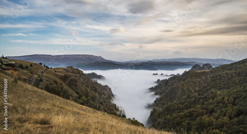 Valley in autumn mountains with clouds below and rocky slopes overgrown with trees and vegetation in autumn, cloudy day in the Caucasus