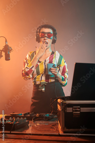 Exhausted performer yawning and feeling sleepy, being tired and drinking coffee after mixing techno song. Musician enjoying beverage or drink and doing stereo remix performance. photo