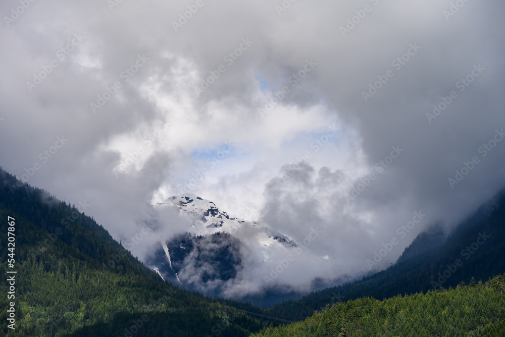 Distant mountain peak peaking out through clouds, forested hills in the foreground
