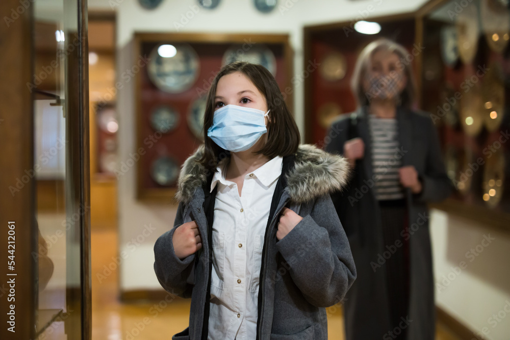 Positive tween schoolgirl in medical face mask observing with interest arts and crafts on exhibition in art gallery. New normal in coronavirus pandemic