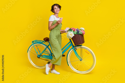 Full size photo of nice young woman hold device bicycle walking flower basket wear trendy khaki outfit isolated on yellow color background © deagreez