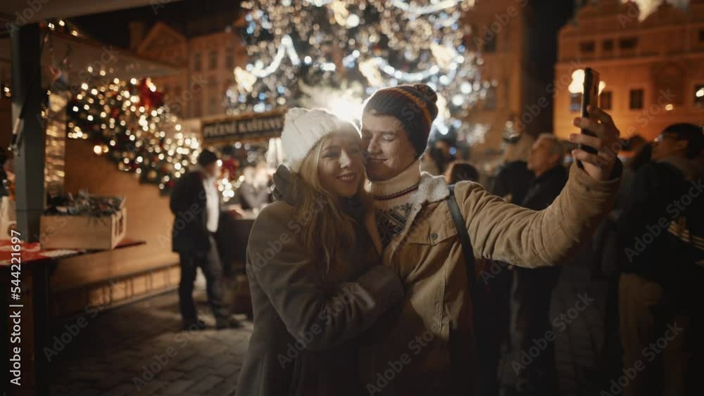 Happy young couple having date at Old decorated town square. Happy time together, People in love celebrate New Year holidays vacation. Woman and man enjoy Christmas, take selie on smartphone and kiss Happy young couple having date at Old decorated town square. Happy time together, People in love celebrate New Year holidays vacation. Woman and man enjoy Christmas, take selie on smartphone and kiss
