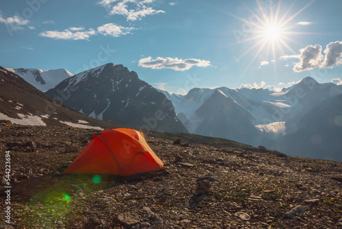 Awesome alpine landscape with orange tent with view to high snow mountains and large glacier. Bright sun in cloudy sky above snowy mountain range. Tent with top view to glacier tongue shining in sun. photo