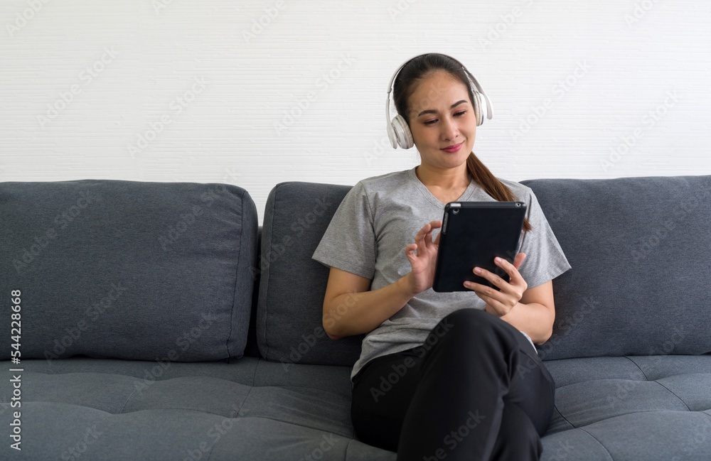 Asian middle-aged woman in gray t-shirt listen to music from headphones while holding tablet computer in the living room, enjoy leisure weekend at home. Stress free concept Asian middle-aged woman in gray t-shirt listen to music from headphones while holding tablet computer in the living room, enjoy leisure weekend at home. Stress free concept