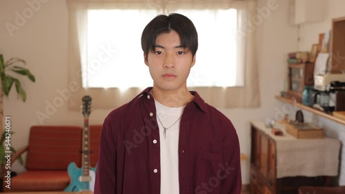 Young asian man stands still with expressionless face at living room. Medium shot.