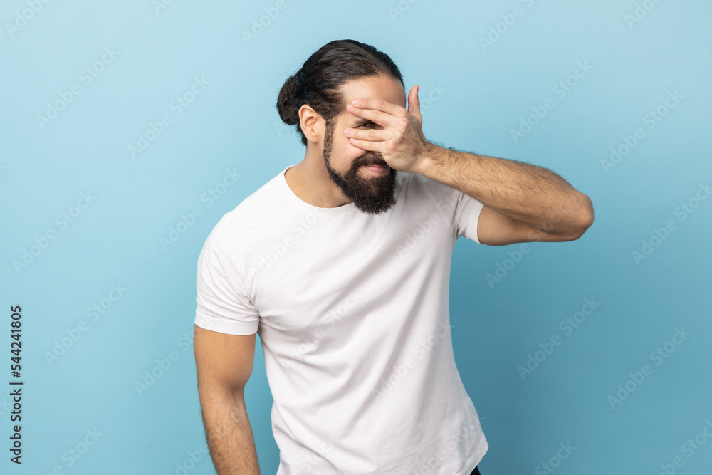 Portrait of man with beard wearing white T-shirt looking through fingers, peeking with curious face, having suspicious, watching secrets. Indoor studio shot isolated on blue background. Portrait of man with beard wearing white T-shirt looking through fingers, peeking with curious face, having suspicious, watching secrets. Indoor studio shot isolated on blue background.