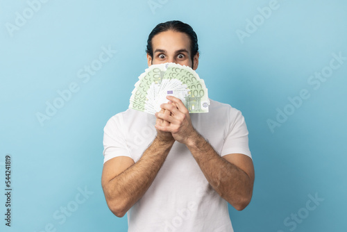 Man with beard wearing white T-shirt covering face with money and peeking out of euro, excited to hold lot of cash, shocked by big lottery win. Indoor studio shot isolated on blue background.